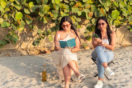 Amigos con libro y smartphone en la playa al atardecer.
