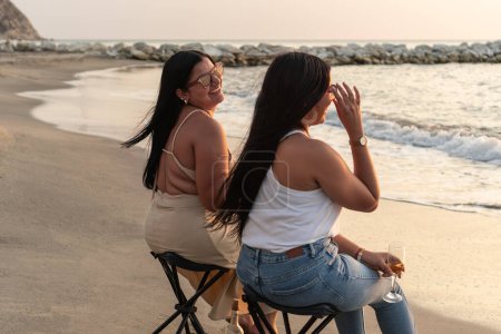 Women laughing and enjoying wine by the ocean at sunset.