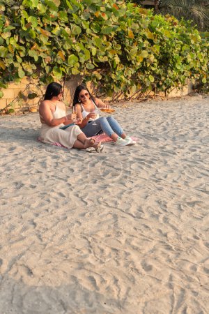 Two women with wine glasses on a beach picnic blanket.