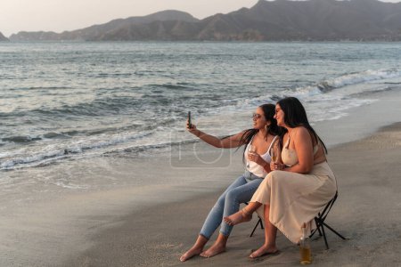 Zwei gut gelaunte Frauen halten einen Selfie-Moment fest, während sie in der Abenddämmerung ein Glas Wein an einem Sandstrand genießen.