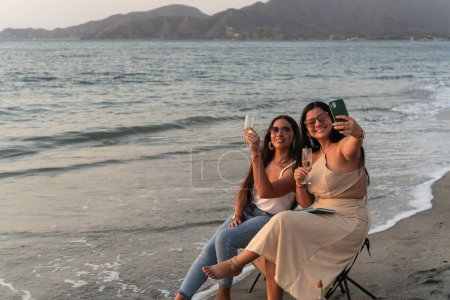Two smiling women taking a selfie while toasting with champagne on a beautiful beach at dusk.