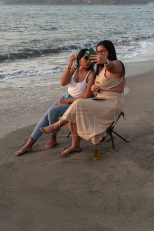 Zwei Frauen posieren für ein verspieltes Selfie, während sie in der Dämmerung mit Wein am Sandstrand anstoßen.