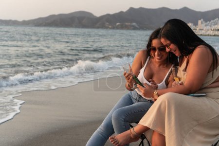 Zwei Frauen lachen, während sie ein Smartphone checken und in der Abenddämmerung Wein an einem malerischen Strand genießen.
