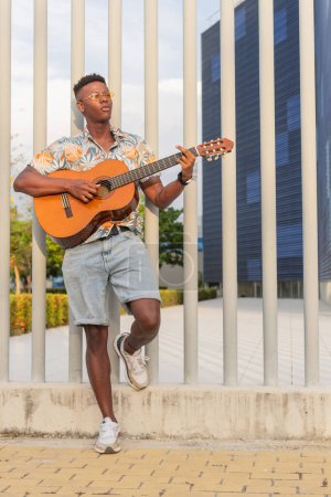 Vertikales Bild eines modischen jungen Afrikaners, der in einem städtischen Park Gitarre spielt, mit stilvoller Sommerkleidung und architektonischem Hintergrund.
