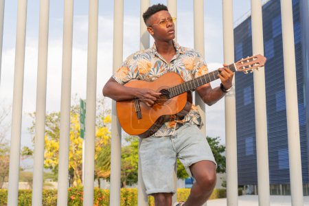 Konzentrierter afrikanischer Mann, der im Park Gitarre spielt, gestylt in einem floralen Hemd und Jeanshosen, mit urbaner Architektur und grünem Hintergrund.