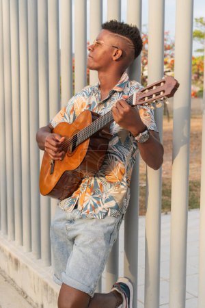 A young African man leans against a metal fence, lost in the music as he plays his guitar, wearing a tropical shirt and sunglasses during sunset.