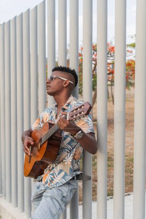 Ein nachdenklicher junger Mann spielt seine Gitarre inmitten einer urbanen Kulisse, umrahmt von lebendiger Flora.