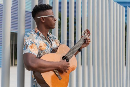 Confident young African American male playing an acoustic guitar against a patterned urban backdrop.