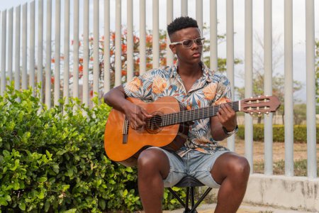 A focused young man wearing a tropical shirt strums an acoustic guitar in a serene outdoor setting with lush greenery and modern architecture.