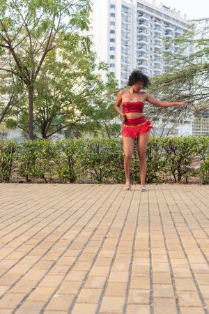 A woman in a vibrant red dance costume poses gracefully on a paved path with lush greenery and tall buildings in the background, showcasing urban and natural elements.