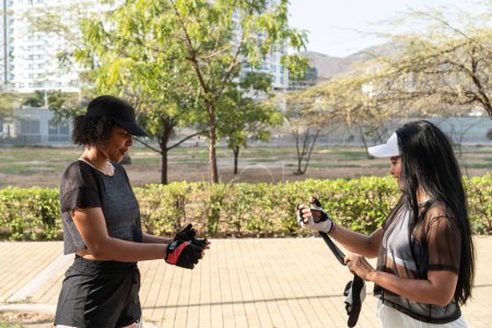 Zwei Frauen, die sich in Sportkleidung und Handschuhen auf ein Workout in einem Park vorbereiten, im Hintergrund Bäume und städtische Gebäude.