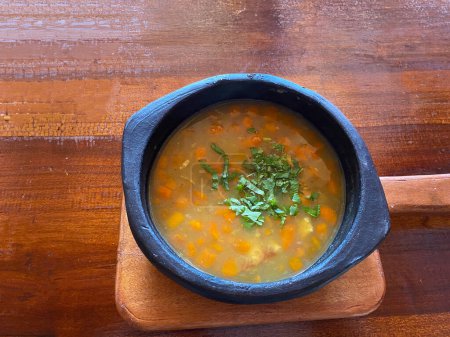 Blick auf eine herzhafte Gemüsesuppe, garniert mit frischen Kräutern, serviert in einer rustikalen schwarzen Steingutschüssel auf einem Holztisch.