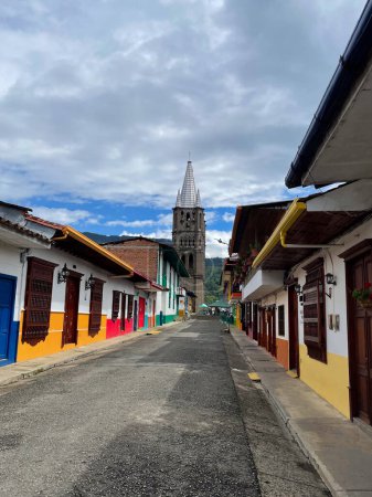 A picturesque view of a colorful colonial street leading to a tall church tower under a cloudy sky, showcasing vibrant buildings and historic architecture.