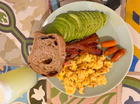 Vue aérienne d'une assiette de petit déjeuner avec des ?ufs brouillés, des tranches d'avocat, du bacon, des saucisses et du pain, servi avec un verre de jus sur une table carrelée colorée.