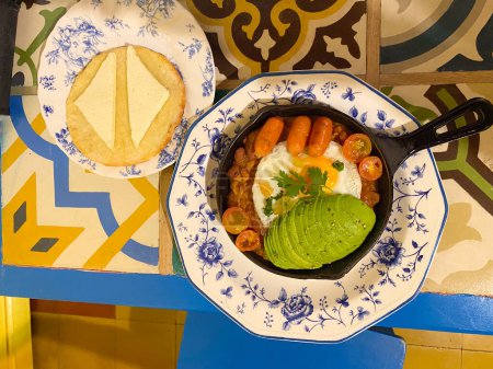 Blick auf ein traditionelles Frühstück mit Spiegelei, Avocadoscheiben, Würstchen und Kirschtomaten in einer Pfanne, serviert mit einem Teller Käse auf Brot.