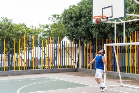 A man in blue sportswear makes a shot on an outdoor basketball court. The ball is just above the hoop, and a colorful fence is in the background