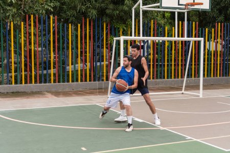 Two basketball players on an outdoor court, one in a defensive stance while the other holds the ball. Colorful poles and green foliage in the background.