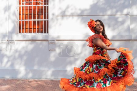 Woman in vibrant traditional dress dancing outdoors on a sunny day with white wall background.