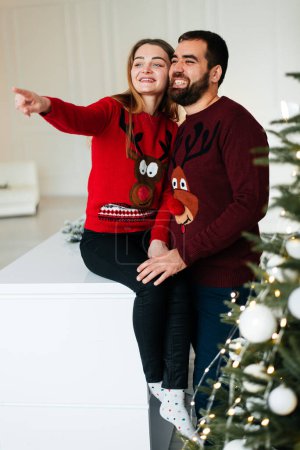 Photo for A couple in love hug and kiss near the Christmas tree in the decorated interior of the Christmas tree on a Christmas holiday in winter at home. A man and a woman celebrate the New year together - Royalty Free Image