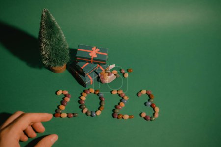 Green Christmas background with decorative pine tree, gifts boxes and female hand. Merry Christmas and Happy New Year Greeting Card with word 2023 made of colorful candies. Winter holidays wallpaper.