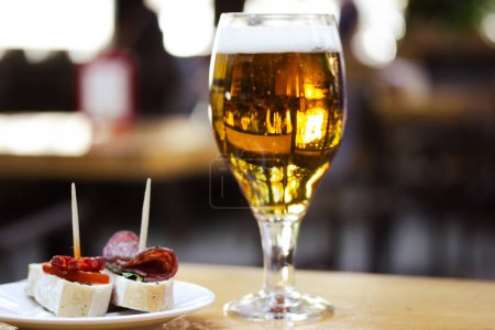 Amber beer in glass. Elegant tableware with golden freshly brewed wheat beer stands on wooden table in cafe, bar pub terrace. Alcoholic beverage with traditional Spanish dish tapas, sausage sandwiches