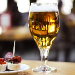 Amber beer in glass. Elegant tableware with golden freshly brewed wheat beer stands on wooden table in cafe, bar pub terrace. Alcoholic beverage with traditional Spanish dish tapas, sausage sandwiches