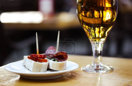 Photo for Amber beer in glass. Elegant tableware with golden freshly brewed wheat beer stands on wooden table in cafe, bar pub terrace. Alcoholic beverage with traditional Spanish dish tapas, sausage sandwiches - Royalty Free Image