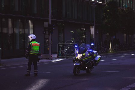 Foto de Berlín, Alemania - 1 de octubre de 2022: Un policía con casco, chaqueta con Polizei escrito en ella patrullando en una calle oscura junto a una motocicleta moderna. Aplicación de la ley en las ciudades. Incidente delictivo - Imagen libre de derechos