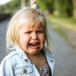 A little preschool-aged 3-5 year old girl is desperately crying loudly standing on a street. The child is lost. Negative emotions. White child refugee from Ukraine. Unhappy toddler kid from orphanage.
