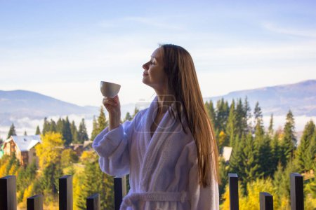 Photo for Young beautiful brunette girl in bathrobe holding cup of tea or morning coffee drink standing on balcony, breathing in clean morning air. Hills, forest, mountains view. Spa recreation. Nature outdoors - Royalty Free Image