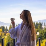 Young beautiful brunette girl in bathrobe holding cup of tea or morning coffee drink standing on balcony, breathing in clean morning air. Hills, forest, mountains view. Spa recreation. Nature outdoors