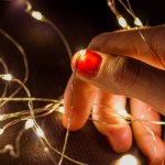 Female hand holding a tangled led garland, glowing lights. Woman preparing to decorate a Christmas tree for New Year's Eve 2023. Thin fingers with orange-red manicure holding a wire on dark background
