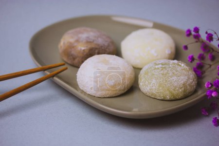 Traditional Japanese dessert mochi in rice dough or daifuku. Mochi ice cream balls and gray round plate, food chopsticks on blue background flatly. Table setting, violet flowers decor. Selective focus