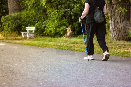 Photo for Nordic walking with sticks, a type of physical activity for retirees. An elderly middle-aged woman does Scandinavian walking outside in a summer park on nature. Sporty active faceless woman back view. - Royalty Free Image