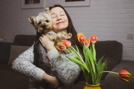 Photo for A young beautiful happy woman cuddling beloved little dog Yorkshire Terrier in a cozy home. A purebred brown puppy pup, a gift for a girl. Sincere emotions of happiness. Orange yellow tulips indoors. - Royalty Free Image