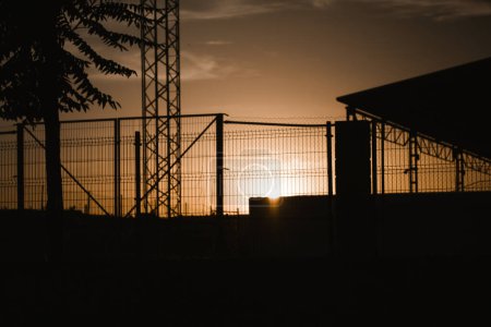 Téléchargez les photos : Contrast silhouette of an industrial building and lattice fence against the sunset yellow brown sky. Warehouse, factory building roof. Commercial property exterior. Wire fence in industrial zone. - en image libre de droit