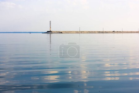 Téléchargez les photos : Lighthouse glowing in a distance. A concrete breakwater on a horizon. A beautiful seascape. A blue sky and reflection in the water water without waves at daytime. Anti-wave protection. Calmness relax. - en image libre de droit