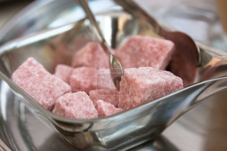 Photo for The pink unrefined lump sugar cubes and the metal sugar serving tongs in a silver sugar bowl on a table in cafe, restaurant, on a kitchen. Sweet unhealthy food. Soft focus. - Royalty Free Image