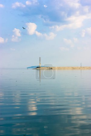 Téléchargez les photos : Lighthouse glowing in a distance. A concrete breakwater on a horizon. A beautiful seascape. A blue sky and reflection in the water water without waves at daytime. Anti-wave protection. Calmness relax. - en image libre de droit