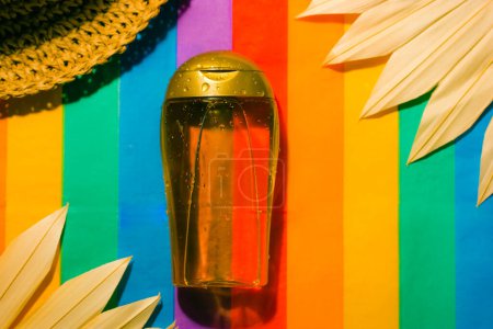 Téléchargez les photos : Suntan oil bottle on multicolored striped bright rainbow background. Sunblock cosmetics. Natural SPF sunscreen product for summer beach vacations. A straw hat, palm leaves. Summer cosmetic still life - en image libre de droit