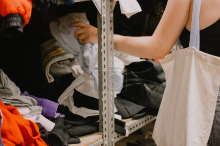 Foto de A young woman at a humanitarian aid station, a flea market, a clothing distribution point for the needy, in a second hand shop choosing shoes among the clothes lying on the shelves Reusability concept - Imagen libre de derechos