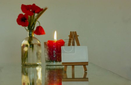 Photo for A wooden miniature easel stands on a table next to a glass vase with red poppies and a red candle with a burning flame. Decor for cozy home. Drawing Lessons, art and hobby concept. Draw a picture. - Royalty Free Image