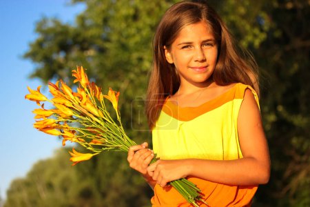Téléchargez les photos : Une belle adolescente heureuse en robe jaune avec un bouquet de lis orange contre un ciel bleu lors d'une journée d'été ensoleillée. Enfant de 10 à 12 ans. 1er septembre, retour à l'école, concept de fête des mères. - en image libre de droit
