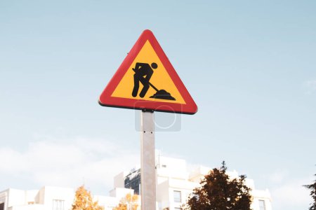 Photo for Roadworks yellow red triangular sign on a city street against blue sky. Temporary construction icon in urban setting. Work in progress. Man is working hard outdoors signboard. Urban landscape. - Royalty Free Image