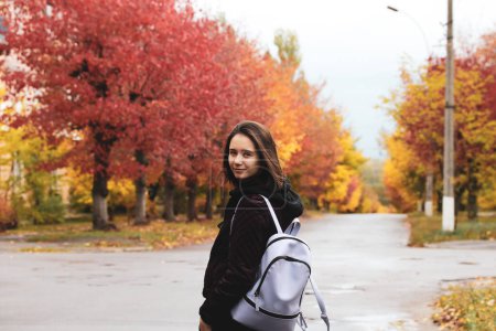 School girl with backpack on a street. Red, yellow trees in fall season. Young woman portrait outdoors in autumnal day. People walking on nature in October, November. Teens on the holidays lifestyle.