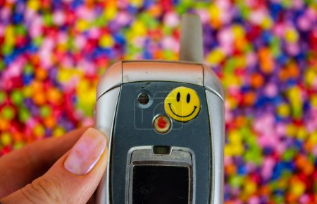 Foto de Madrid, España. Enero 2, 2024 Teléfono Motorola viejo con cara sonriente pegatina en la mano de la mujer. Nostalgia de los 90, 00. Mano sosteniendo teléfono vintage. - Imagen libre de derechos