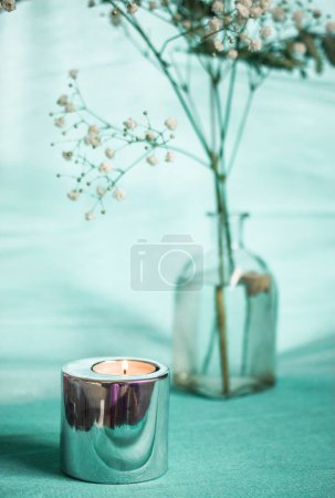 A burning candle in metal candle holder on blue table. Stylish decor for home interior. Still life with a glass vase and small white blooming flowers.