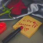 Red lipstick and hand written note with text Love yourself, heart shaped draw on cosmetic table. A single rose, heart candle on Saint Valentines Day. 