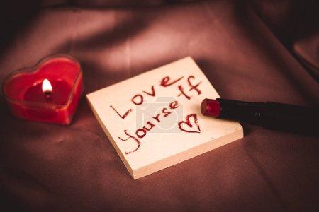 A note to self with a call to love yourself drawing with red lipstick on a silk dark pink cloth flatly. A burning heart-shaped red candle, gift box. Happy Valentines Day 2024. Love yourself first.