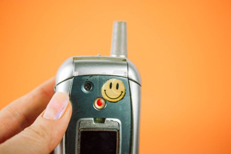 Foto de Madrid, España. Enero 2, 2024 Teléfono Motorola viejo con cara sonriente pegatina en la mano de la mujer. Nostalgia de los 90, 00. Mano sosteniendo teléfono vintage. - Imagen libre de derechos